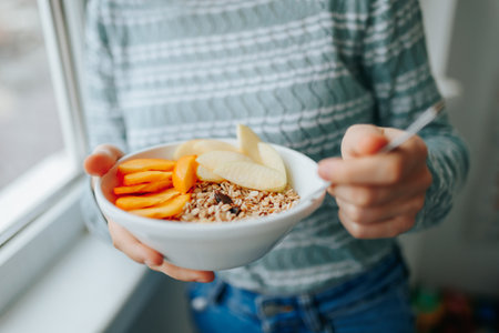 Granola, seeds, fresh fruits in a white bowl in female hands with spoon, unrecognizable. Homemade granola or muesli with oat flakes, corn flakes, dried fruits with fresh apple. Healthy Breakfastの写真素材