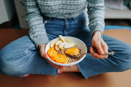 Unrecognizable woman sits at home on floor in Turkish pose holding plate and spoon with delicious breakfast of plant based lean foods. Dietary food, healthy food, fruit on spoon, breakfast plate.の写真素材