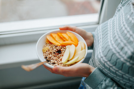 Unrecognizable top view woman in home clothes stands at the window and holds a full plate of granola muesli in her hands. Complete nutritious breakfast for health conscious people.の写真素材