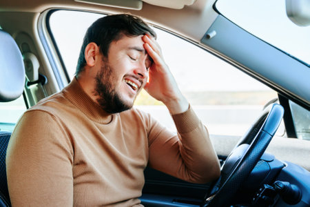 A young man sits behind the wheel in a car with his eyes closed, smiling tiredly, rubbing his forehead with his hand. The road was long and difficult. The guy is resting. People and emotions.の写真素材