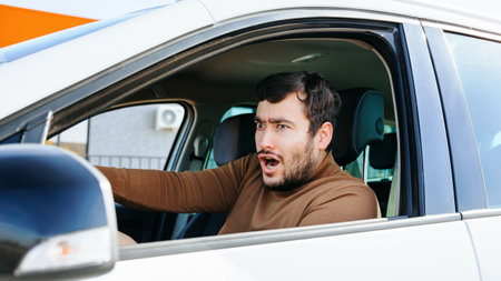 Front view of a young guy, a man grimaces while sitting in a car, apparently something excited him. The driver behind the wheel in a beige sweater with a blue collar. Human emotions and people.の写真素材