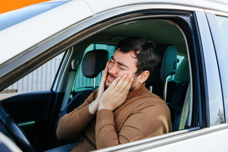 The driver of the car is a middle aged man suffering from a toothache while sitting in a vehicle. Keeps hands on face and winces. The hard work of a car driver.の写真素材
