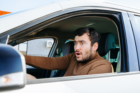 A young man sits in a car, looks in the side mirror and is surprised and frightened by something. Maybe there was a long traffic jam that seriously slowed down the movement. Man, human feelings.の写真素材