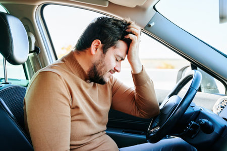A tired young middle aged man sits in a car with his hand on his head with his eyes closed. Bad mood and well being do not allow to drive normally. People and emotions.の写真素材