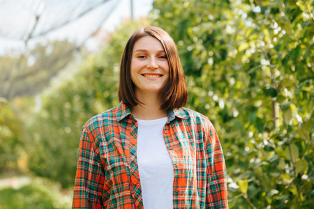 Front view portrait young female farmer girl short hair orchard stands smiling broadly. Female satisfied with the work done, dressed in a plaid shirt and a white T-shirt. Farming concept harvesting.の写真素材