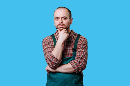 contemplative young workman thoughtfulness in this studio portrait. Dressed in overalls and captured against a stunning blue background, professional deep in thought as he gazes off into the distance.の写真素材
