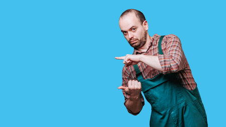 wondering handsome man dressed in a coverall is captured in an unusual pose deep thought, staring intently looking at camera and pointing with his hand. copy space, studio shot, blue backgroundの写真素材