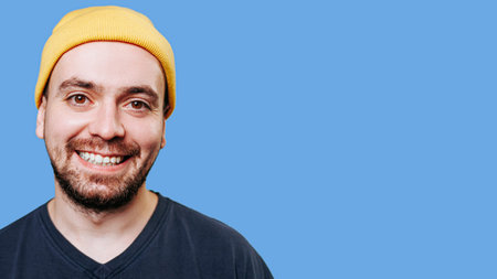 A man in a yellow cap, with a wide and beaming smile on his face, exuding an air of joy and satisfaction, in a studio shot against a blue background, looking intently at the camera with copy space.の写真素材