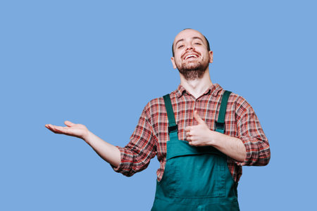 The smiling face of the guy is matched by the positivity conveyed by his outstretched hand giving a thumbs up, against a blue studio background. copy space, Pleased, contented man dressed in coverallの写真素材