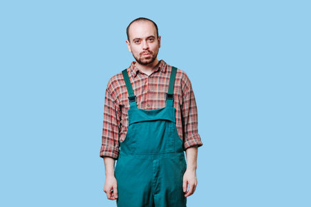 A young, bearded workman pulls funny faces and looks directly at the camera while dressed in a work outfit, set against a blue studio background. bearded farmer makes silly expressions. copy spaceの写真素材