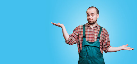 A wandering guy with spreading hands who is wearing a coverall and expressing a curious and inquisitive emotion as he gazes into the camera. studio shot on the blue background, copy spaceの写真素材