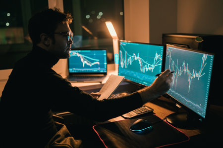 Analyzing the Market In a dimly lit home office, a male trader reviews charts and indexes, illuminated only by the glow of his multi-screen setup.の写真素材