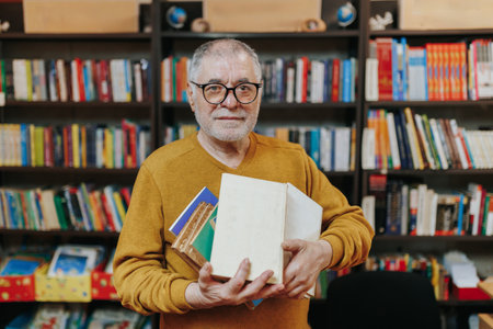 A senior man with a distinguished beard and glasses holds a stack of books in his hand, browsing through the shelves of a bookstore.の写真素材