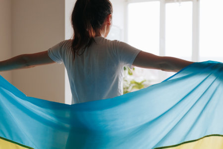 By the window in her home, a womans back faces the camera as she holds the Ukrainian flag, expressing her allegiance to her country.の写真素材