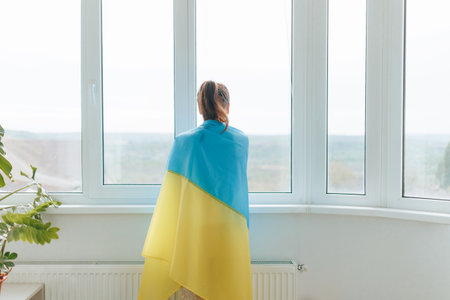 From behind, a woman stands near the window at home, holding the Ukrainian flag in her hand, evoking a sense of patriotism.の写真素材