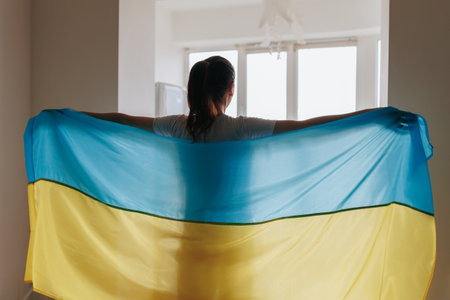 In a conceptual image, a womans rear view depicts her holding the Ukrainian flag by the window in her home, resonating with national pride.の写真素材