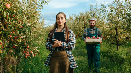 Front view in an apple garden three farmers woman stands smiling tablet in hands looks into the camera men harvest. Young farmers field workers with mustache beard in work clothes. Family business.の写真素材