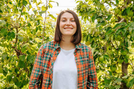 Looking at camera front view stand in orchard of fruit trees young farmer woman smiling joyful. Agronomy adult girl is glad harvest rich. Family garden, family business, young business is successful.の写真素材