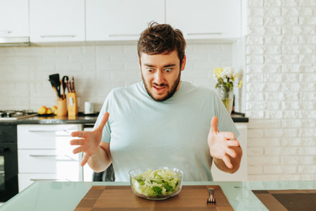 A young man sits in the kitchen in front of a glass bowl of salad with mixed feelings. He looks at the salad with bulging eyes. Greens again!の写真素材