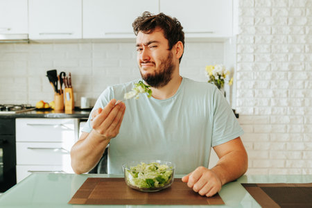 Frustrated by the need to eat unloved food, a young guy holds a fork with a green salad in his hand. Diet food is not what the guy dreamed of. Stop diet, healthy food, vegan food.の写真素材
