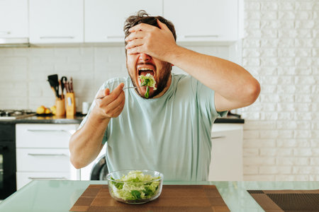 With mouth wide open young guy man brings fork with salad to his mouth, closing his eyes with his hand. Healthy diet, raw vegetables and greens. It is useful to monitor health and weight. Stop diet.の写真素材