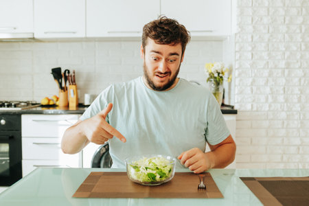 Sitting in kitchen looking into a bowl of salad a man thinks how tired he is of healthy lifestyle. Young man wants to be healthy, but its hard and not fun. Curves faces, showing different emotions.の写真素材