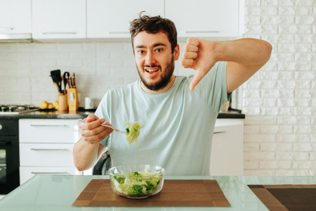Sadly showing a dislike, a young guy holds a fork with a green juicy salad in his hand. The diet is useful, but it spoils the mood. Stop dieting. It is time to improve your mood.の写真素材