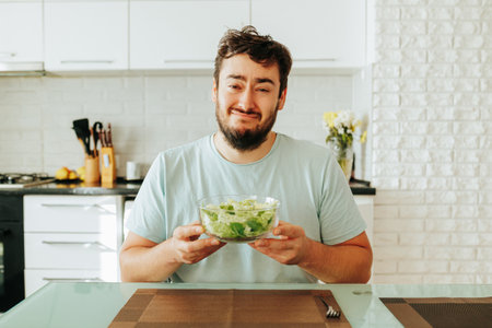 A young man with a martyred expression looking at the camera and holds a glass bowl of salad in his hands. The guy suffers from the need to eat vegetables and salad. Stop diet. Front view.の写真素材