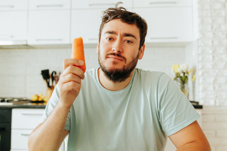Front view looking at the camera a young man indifferently and slightly tormented holds a raw carrot in his hand. He must be on the right diet. This puts him in a bad mood.の写真素材