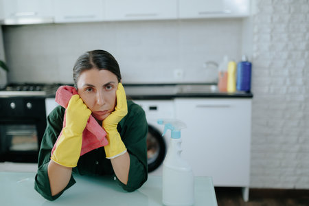 Exhausted Woman in Kitchen, Seated at Table with Yellow Glovesの写真素材