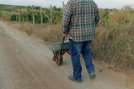 Elderly Farmer Walking with Wheelbarrow on Country Pathの写真素材