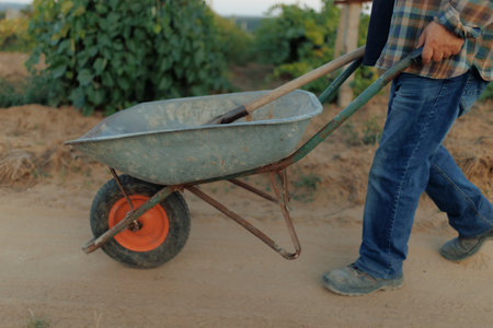 Close-Up Old Farmer Pushing Wheelbarrow Along Country Roadの写真素材