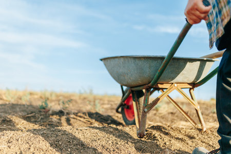 Close-Up of an Elderly Farmer Pushing Wheelbarrow on Dirt Road Near Fieldの写真素材