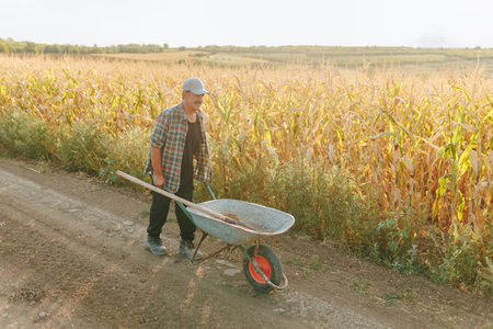 Senior Farmer Pushing Wheelbarrow on Country Road Near Fieldの写真素材
