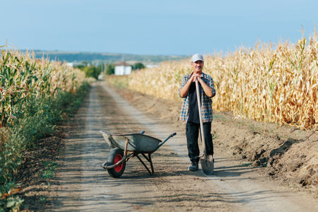 Senior Farmer with Cap and Shovel Poses by Wheelbarrow on Country Roadの写真素材