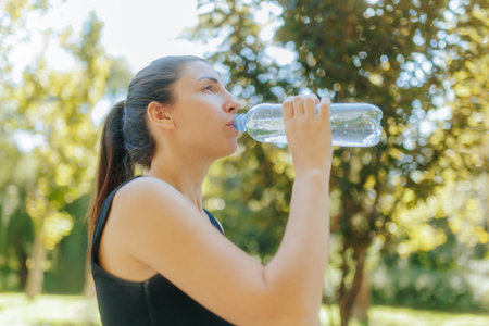 Young white woman drinks water from a bottle after fitness training outdoors in the park