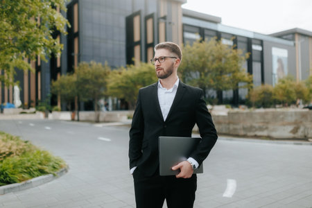 Serious Bearded Man with Glasses Holding Laptop Outside Office Buildingの写真素材
