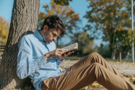 Astonished Man Reads Book Under a Tree, Fall Leaves Surrounding Himの写真素材