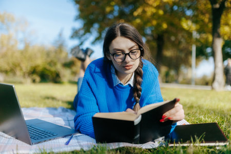 A teenage girl with glasses sits on the grass with a book, studying and reading in a calm autumn park. A laptop beside her reflects a blend of nature and technology.の写真素材