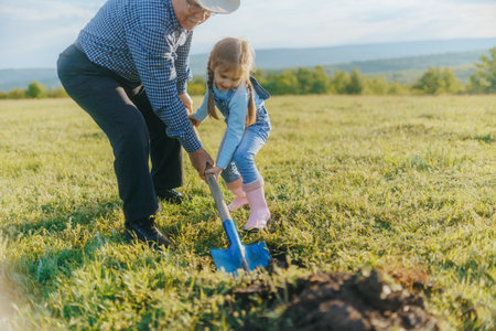 A grandfather in a cowboy hat and his granddaughter dig a hole with a shovel, preparing to plant a tree in the quiet countryside during a golden sunset.の写真素材