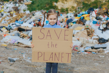 A young girl stands in front of a landfill holding a cardboard sign that reads Save the Planet, calling attention to environmental issues and the need for cleaner habits.の写真素材