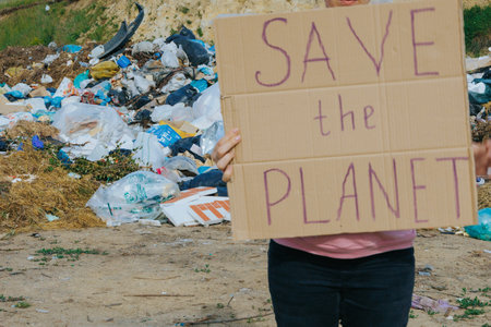 A female activist holds a cardboard sign reading SAVE THE PLANET in front of a polluted landfill, urging for climate awareness, recycling, and environmental change.の写真素材
