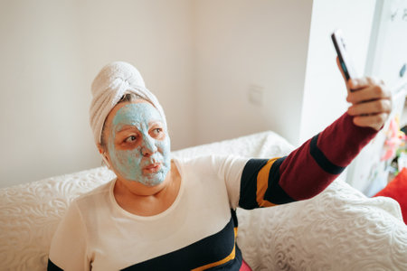 Woman with facial mask taking selfie at home during relaxation timeの写真素材