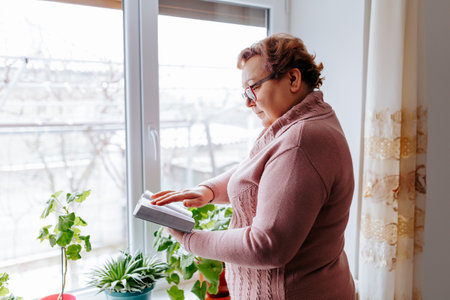 Senior woman reading book while standing near window with plants indoorsの写真素材