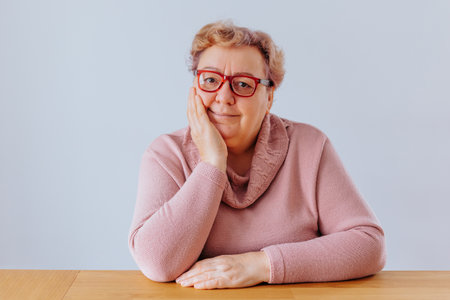 Elderly woman in cozy sweater sitting at table with thoughtful expressionの写真素材
