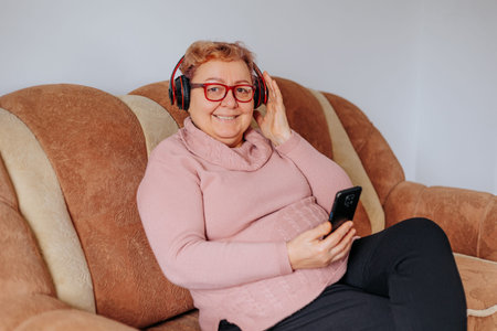 An older woman sits on a comfortable couch, wearing headphones and smiling