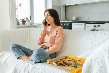 Young woman enjoys pizza while talking on the phone in cozy kitchen settingの写真素材