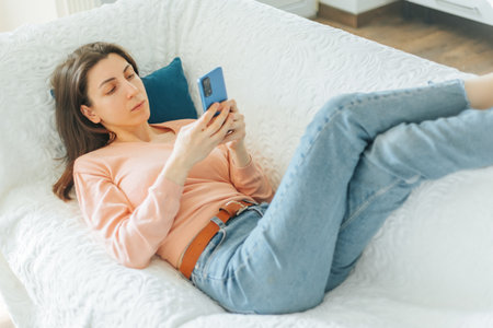 Woman relaxing on a sofa while using a smartphone in a cozy living roomの写真素材