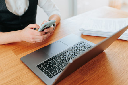Office worker using smartphone while sitting at wooden desk with laptop