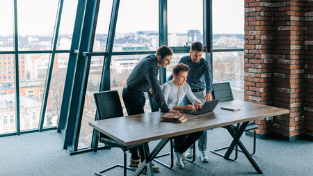 Colleagues collaborating around a laptop in a modern office for brainstorming ideas and data...